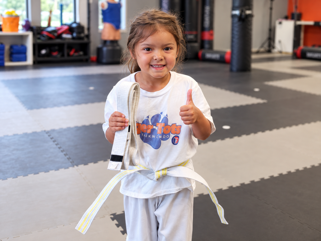 Smiling Tiger Tots student giving a thumbs up while holding her belt in the dojang.
