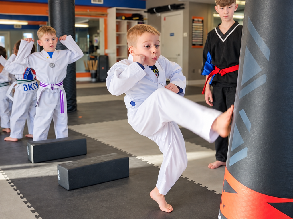 Tiger Tots student practicing a front kick on a punching bag with an instructor nearby.